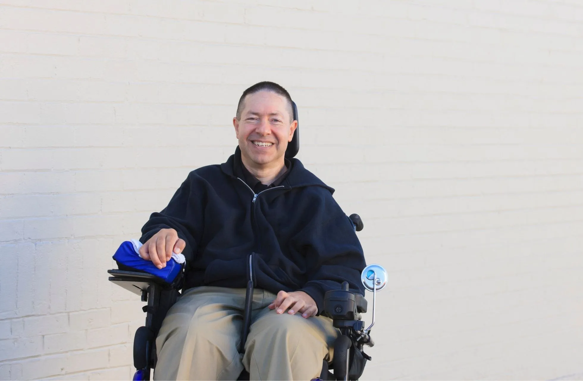 A white man with short dark hair and shaved sides is sat in his wheelchair, in front of a white painted brick wall. He is smiling, and is wearing khaki trousers, and a zipped up black jacket.
