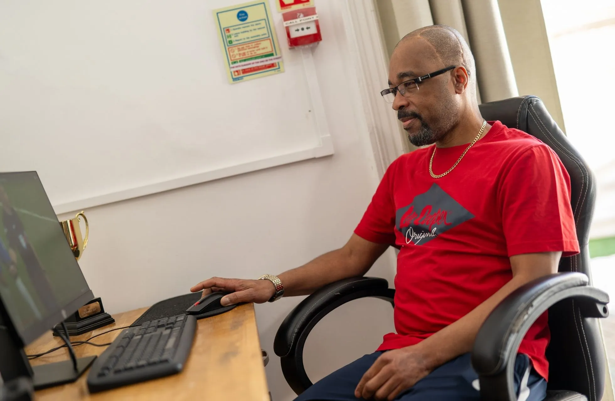 An Active Assistance client is sat on a black desk chair at a wooden desk, using a computer. He is a black man with eye glasses, a short beard, gold necklace and red t-shirt.