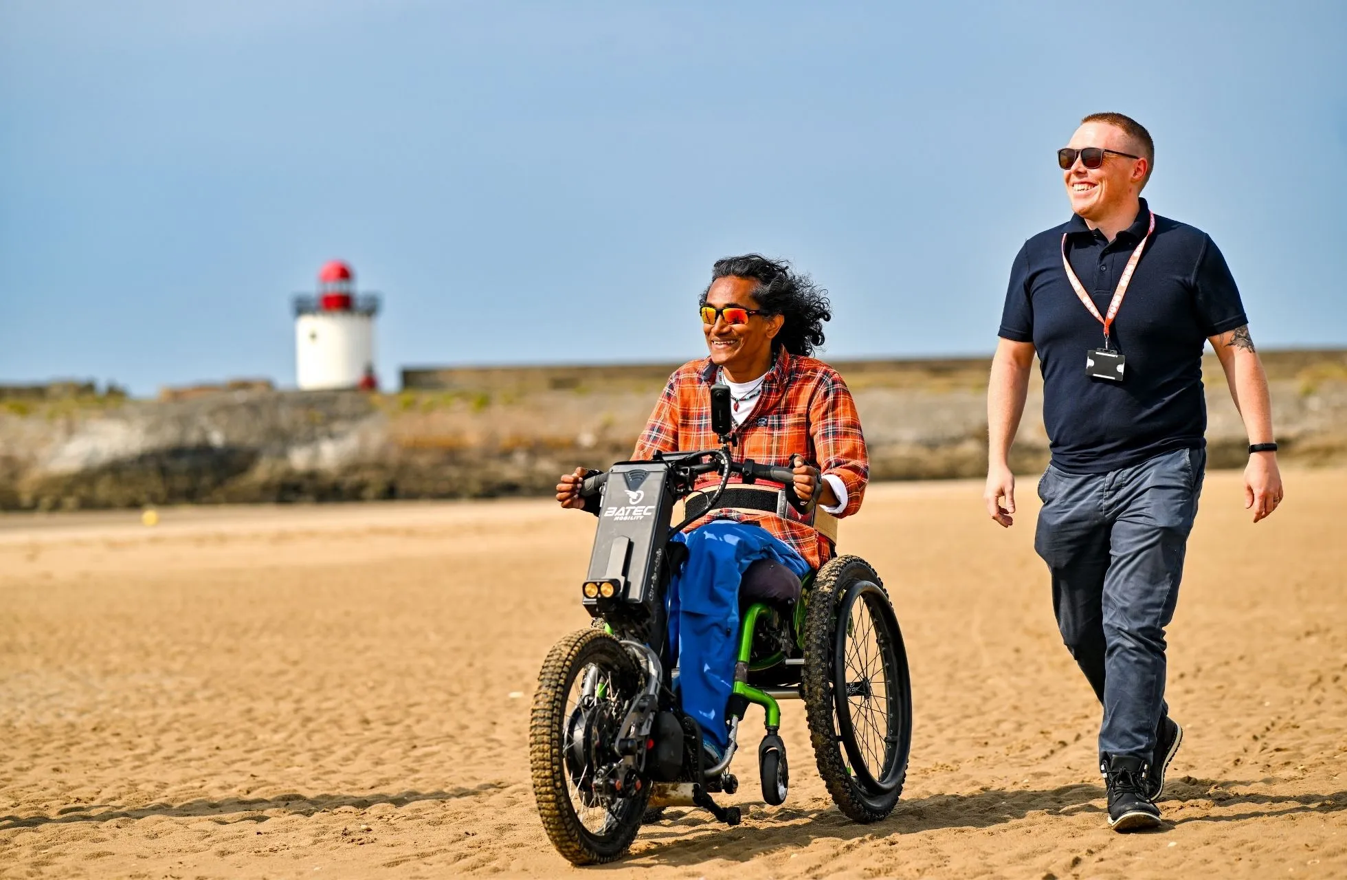Active Assistance client, Kevin, rides an adapted motorbike on a sandy beach, with his support worker standing next to him. They are both smiling.